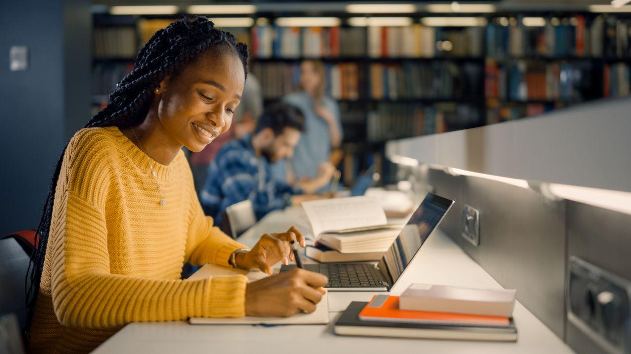 Laechelnde Studentin schreibt in der Bibliothek konzentriert an ihrer Arbeit und nutzt Planung fuer Bachelorarbeiten schreiben lassen